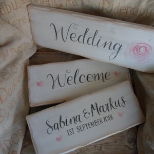 Pu&ograve; includere: Tre cartelli rustici in legno con superfici verniciate di bianco. Il cartello superiore recita "Wedding" con un'illustrazione di rosa rosa. Il secondo cartello dice "Welcome" con un cuore rosa. Il cartello inferiore recita "Sabina & Markus 1st September 2018".