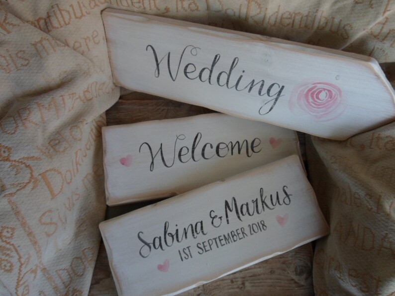 Pu&ograve; includere: Tre cartelli rustici in legno con superfici verniciate di bianco. Il cartello superiore recita "Wedding" con un'illustrazione di rosa rosa. Il secondo cartello dice "Welcome" con un cuore rosa. Il cartello inferiore recita "Sabina & Markus 1st September 2018".