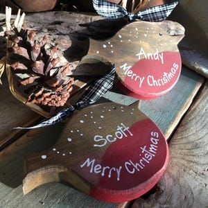 May include: Two wooden bird-shaped ornaments featuring a brown top, a red breast, and white painted details. One ornament is personalized with the name Andy and the other with Scott, both inscribed with the text Merry Christmas and accented with black and white checkered ribbons.