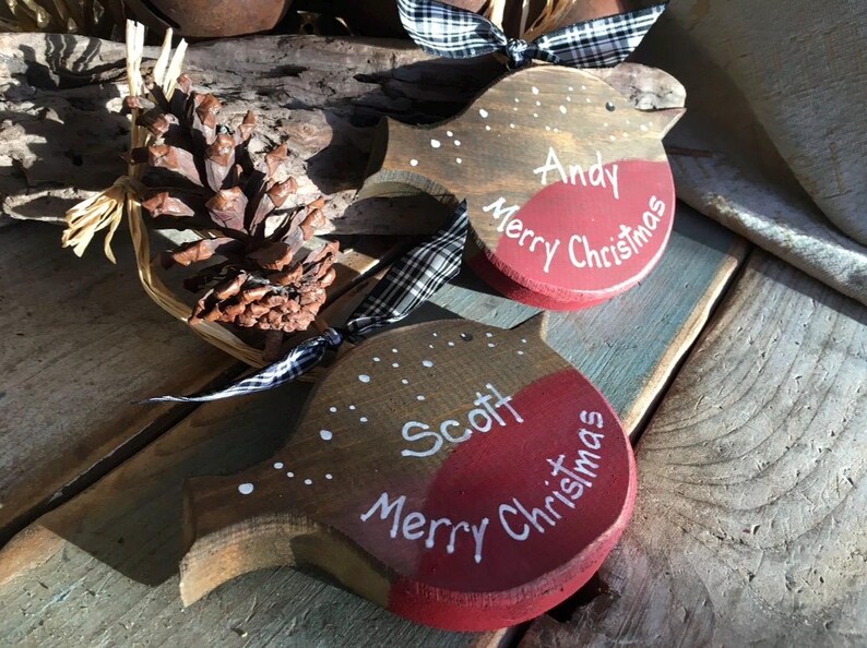 May include: Two wooden bird-shaped ornaments featuring a brown top, a red breast, and white painted details. One ornament is personalized with the name Andy and the other with Scott, both inscribed with the text Merry Christmas and accented with black and white checkered ribbons.