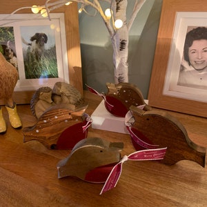 May include: Four wooden robin ornaments with brown stained bodies and red painted chests. Each bird features a small black eye and a ribbon tied around its middle that reads Merry Christmas in white script. The ornaments are arranged on a wooden surface.