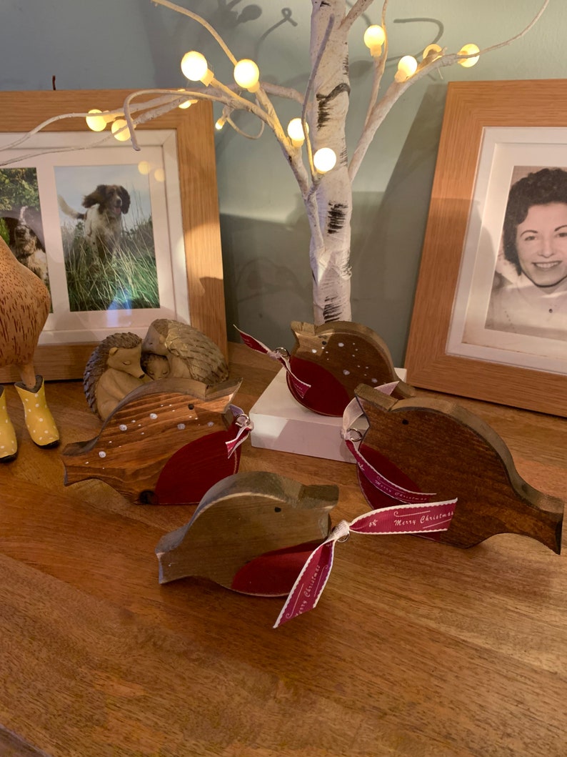 May include: Four wooden robin ornaments with brown stained bodies and red painted chests. Each bird features a small black eye and a ribbon tied around its middle that reads Merry Christmas in white script. The ornaments are arranged on a wooden surface.