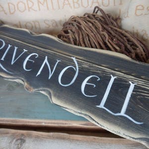 Pu&ograve; includere: Un cartello in legno invecchiato con le parole "Rivendell" dipinte in bianco. Il cartello &egrave; marrone scuro e ha un aspetto rustico e invecchiato.