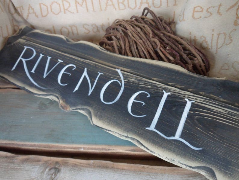 Pu&ograve; includere: Un cartello in legno invecchiato con le parole "Rivendell" dipinte in bianco. Il cartello &egrave; marrone scuro e ha un aspetto rustico e invecchiato.