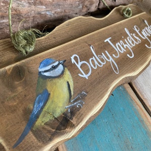 K&ouml;nnte beinhalten: Ein Holzschild mit dem Text "Baby Jazzie's Garden" in wei&szlig;er Farbe. Auf der linken Seite des Schildes ist ein Blaumeise gemalt.