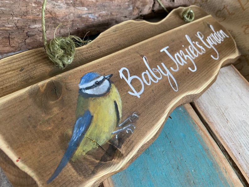 K&ouml;nnte beinhalten: Ein Holzschild mit dem Text "Baby Jazzie's Garden" in wei&szlig;er Farbe. Auf der linken Seite des Schildes ist ein Blaumeise gemalt.