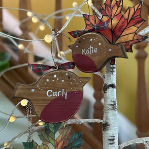 May include: Two wooden robin ornaments with red chests and brown bodies, each featuring a white name, 'Carly' and 'Katie', and a small plaid ribbon. They are displayed alongside two stained glass maple leaf ornaments in autumn colors.