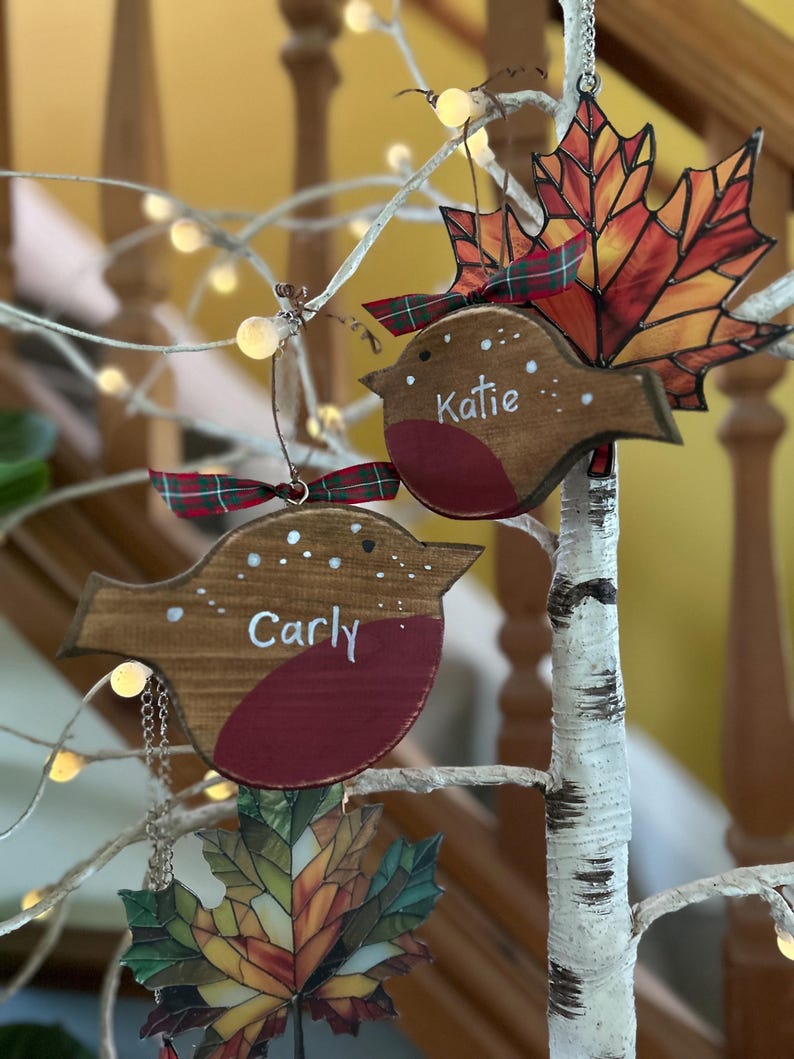 May include: Two wooden robin ornaments with red chests and brown bodies, each featuring a white name, 'Carly' and 'Katie', and a small plaid ribbon. They are displayed alongside two stained glass maple leaf ornaments in autumn colors.