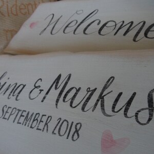 Pu&ograve; includere: Due cartelli in legno bianco con calligrafia nera. Il cartello in alto recita "Welcome". Il cartello in basso dice "Sabina & Markus 1 settembre 2018" con accenti a forma di cuore rosa. I cartelli hanno un aspetto rustico e fatto a mano.