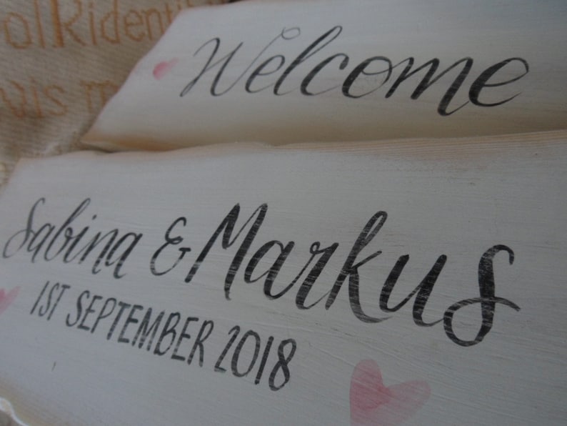Pu&ograve; includere: Due cartelli in legno bianco con calligrafia nera. Il cartello in alto recita "Welcome". Il cartello in basso dice "Sabina & Markus 1 settembre 2018" con accenti a forma di cuore rosa. I cartelli hanno un aspetto rustico e fatto a mano.