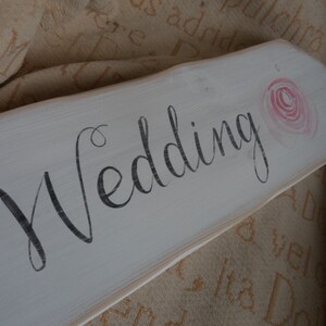 Pu&ograve; includere: Un cartello di legno bianco con la parola "Wedding" in corsivo nero. Una rosa rosa ad acquerello &egrave; dipinta nell'angolo in alto a destra. Il cartello ha un aspetto rustico e invecchiato, ideale per le decorazioni nuziali.