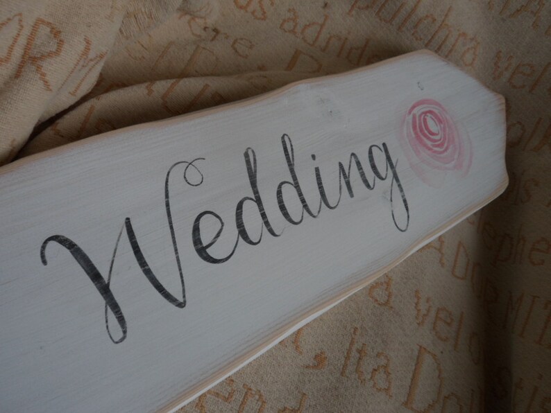 Pu&ograve; includere: Un cartello di legno bianco con la parola "Wedding" in corsivo nero. Una rosa rosa ad acquerello &egrave; dipinta nell'angolo in alto a destra. Il cartello ha un aspetto rustico e invecchiato, ideale per le decorazioni nuziali.