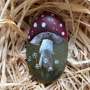 Puede incluir: Una piedra de forma ovalada pintada con un diseño detallado de hongo. El hongo presenta un sombrero rojo oscuro con manchas blancas y un tallo blanco, sobre un fondo verde oliva oscuro con pequeños detalles florales blancos en la base.