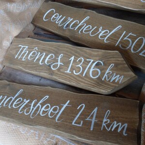 Pu&ograve; includere: Segnali stradali in legno con scritte bianche. I segnali indicano le distanze in chilometri verso varie localit&agrave;: Courchevel 1502 km, Th&ocirc;nes 1376 km e Laundersfoot 24 km. Il legno ha una finitura marrone naturale.