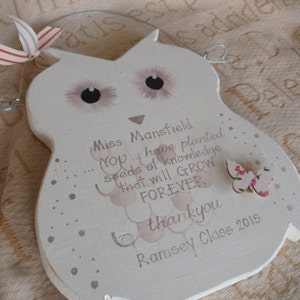 Op de afbeelding: Een wit houten uilvormig bord met een roze vlinder, de tekst "Miss Mansfield You... have planted seeds of knowledge that will GROW FOREVER thankyou Ramsey Class 2015" en een roze en wit stippenpatroon.