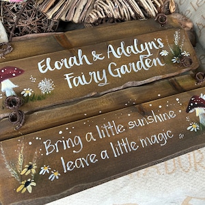 K&ouml;nnte beinhalten: Zwei Holzschildchen mit gemalten Pilzen, Blumen und dem Text "Elorah & Adalyn's Fairy Garden" und "Bring a little sunshine leave a little magic".