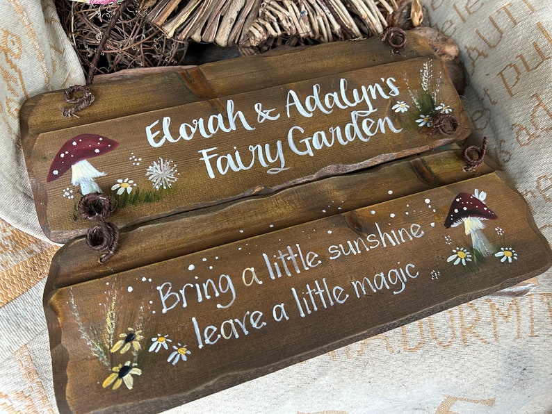 K&ouml;nnte beinhalten: Zwei Holzschildchen mit gemalten Pilzen, Blumen und dem Text "Elorah & Adalyn's Fairy Garden" und "Bring a little sunshine leave a little magic".