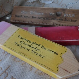May include: Two wooden pencil-shaped signs with painted details. The top sign is brown and says "Miss Williams... YOU RULE thank you... for being an awesome teacher". The bottom sign is yellow and says "teachers plant the seeds of knowledge that grow forever".