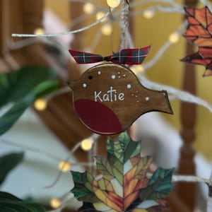 May include: A wooden bird ornament with a brown body, a red chest, and white painted details including the name Katie. The ornament is topped with a small green and red plaid ribbon and hangs from a string, featuring a simple cutout shape.
