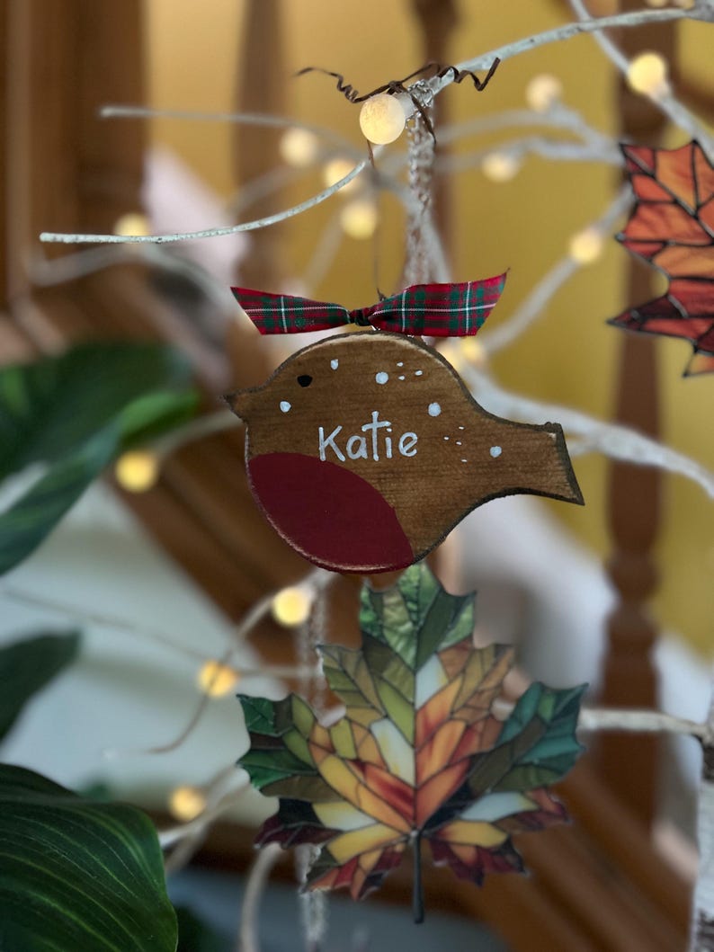 May include: A wooden bird ornament with a brown body, a red chest, and white painted details including the name Katie. The ornament is topped with a small green and red plaid ribbon and hangs from a string, featuring a simple cutout shape.