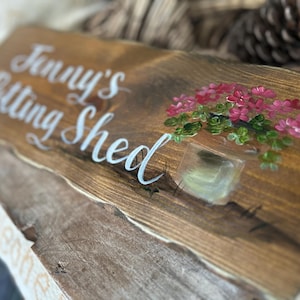 May include: A rectangular wooden sign with a natural live edge, stained in a medium brown tone. The text "Jenny's Potting Shed" is painted in white script, accompanied by a hand-painted illustration of a potted plant with pink flowers.