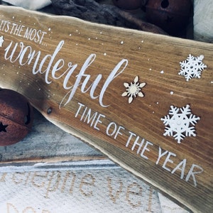 Può includere: Un cartello di legno con il testo "It's the most wonderful time of the year" dipinto di bianco. Il cartello è decorato con fiocchi di neve bianchi e un fiocco di neve dorato.