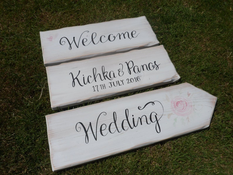Pu&ograve; includere: Tre cartelli nuziali in legno bianco con scritte nere. Il cartello in alto recita "Welcome", il cartello centrale dice "Kichka & Panos 17th July 2016", e il cartello in basso dice "Wedding" con un accento floreale rosa.