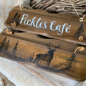 K&ouml;nnte beinhalten: Zwei Holzschildchen mit rustikalem Finish. Das obere Schild tr&auml;gt die Aufschrift "Pickle's Cafe" in wei&szlig;er, kursiv geschriebener Schrift. Das untere Schild zeigt eine Silhouette eines Hirsches in einer Waldlandschaft.