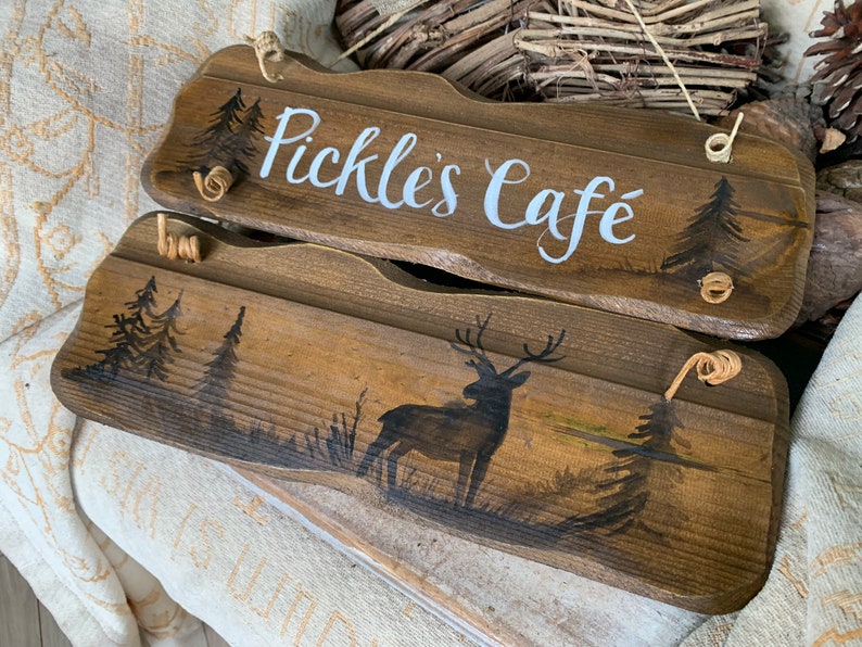 K&ouml;nnte beinhalten: Zwei Holzschildchen mit rustikalem Finish. Das obere Schild tr&auml;gt die Aufschrift "Pickle's Cafe" in wei&szlig;er, kursiv geschriebener Schrift. Das untere Schild zeigt eine Silhouette eines Hirsches in einer Waldlandschaft.