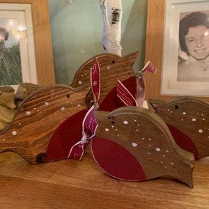May include: Three wooden bird ornaments with brown stained bodies and red painted chests. Each bird features small white painted dots resembling snow and a dark eye. One bird is tied with a red ribbon printed with white snowflakes and text.
