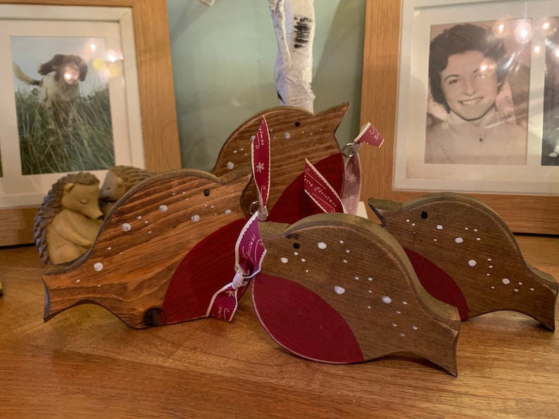 May include: Three wooden bird ornaments with brown stained bodies and red painted chests. Each bird features small white painted dots resembling snow and a dark eye. One bird is tied with a red ribbon printed with white snowflakes and text.
