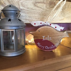 May include: A wooden bird-shaped ornament with a red painted chest, featuring the white text "Isla" and "Merry Christmas". The ornament is adorned with a white ribbon with red polka dots and a natural raffia tie for hanging.