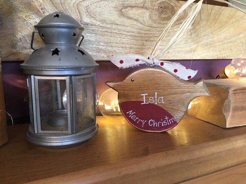 May include: A wooden bird-shaped ornament with a red painted chest, featuring the white text "Isla" and "Merry Christmas". The ornament is adorned with a white ribbon with red polka dots and a natural raffia tie for hanging.