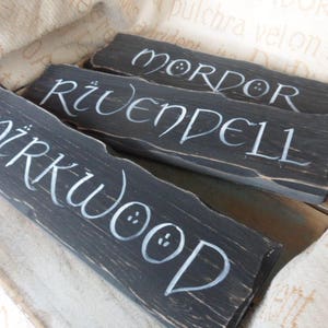 Pu&ograve; includere: Tre cartelli di legno dipinti di nero con lettere bianche. I cartelli recitano "Mirkwood", "Rivendell" e "Mordor".