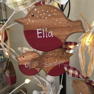 May include: A wooden bird-shaped ornament with a natural wood finish, a painted red chest, and white polka dots on the back. The name Ella is hand-painted in white on the chest. A plaid ribbon is tied to the top for hanging purposes.