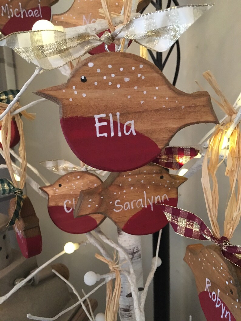 May include: A wooden bird-shaped ornament with a natural wood finish, a painted red chest, and white polka dots on the back. The name Ella is hand-painted in white on the chest. A plaid ribbon is tied to the top for hanging purposes.