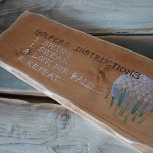 Puede incluir: Un letrero de madera con el texto "Golfers Instructions" y una lista de instrucciones: 1. Swing, 2. Swear, 3. Look for ball, 4. Repeat. El letrero tambi&eacute;n presenta una pelota de golf pintada.