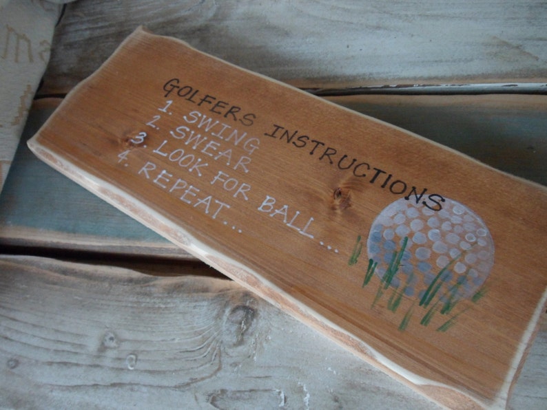 Puede incluir: Un letrero de madera con el texto "Golfers Instructions" y una lista de instrucciones: 1. Swing, 2. Swear, 3. Look for ball, 4. Repeat. El letrero tambi&eacute;n presenta una pelota de golf pintada.