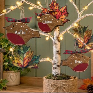 May include: Three wooden bird ornaments with red chests and white names Katie, Chloe, and Evie, hanging from a lit birch tree alongside stained glass-style maple leaf ornaments. Each bird features a small plaid ribbon at the top.
