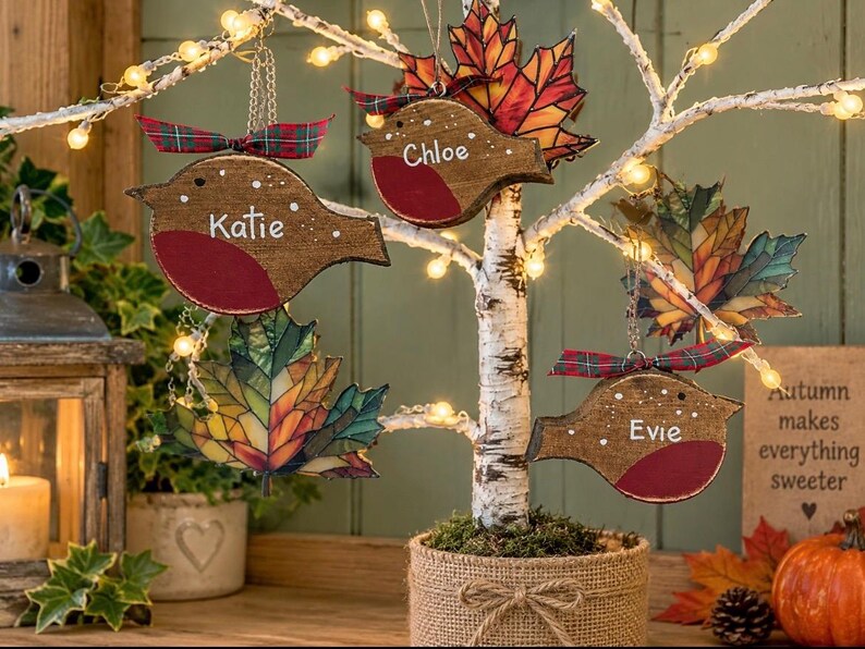 May include: Three wooden bird ornaments with red chests and white names Katie, Chloe, and Evie, hanging from a lit birch tree alongside stained glass-style maple leaf ornaments. Each bird features a small plaid ribbon at the top.