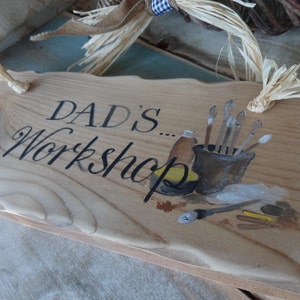 Puede incluir: Un letrero de madera con las palabras "Dad's Workshop" pintadas en letra cursiva negra. El letrero está decorado con una imagen pintada de pinceles en un frasco, una botella de pintura y otros materiales de arte.
