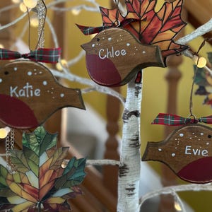 May include: Three wooden robin ornaments with red chests, each personalized with the white painted names Katie, Chloe, and Evie. The birds feature green and red plaid ribbon bows and are displayed alongside stained glass-style maple leaf ornaments.