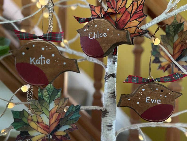 May include: Three wooden robin ornaments with red chests, each personalized with the white painted names Katie, Chloe, and Evie. The birds feature green and red plaid ribbon bows and are displayed alongside stained glass-style maple leaf ornaments.