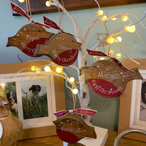 May include: Four wooden robin-shaped Christmas ornaments hanging from a white lighted tree. Each ornament features a red breast, white painted text including names like Popsi, Nanny, and Maisie, and a red ribbon printed with Merry Christmas.