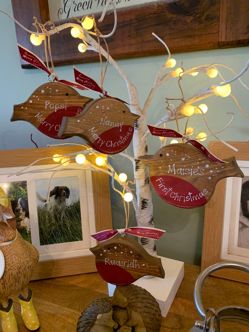 May include: Four wooden robin-shaped Christmas ornaments hanging from a white lighted tree. Each ornament features a red breast, white painted text including names like Popsi, Nanny, and Maisie, and a red ribbon printed with Merry Christmas.