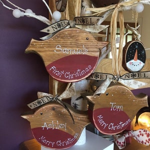 May include: Three wooden bird-shaped Christmas ornaments with natural wood grain, red painted chests, and white lettering. Each ornament features a name and holiday greeting, tied with beige ribbon printed with the word NOEL.