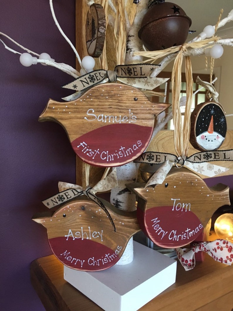 May include: Three wooden bird-shaped Christmas ornaments with natural wood grain, red painted chests, and white lettering. Each ornament features a name and holiday greeting, tied with beige ribbon printed with the word NOEL.