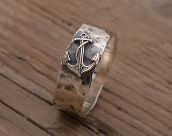Könnte beinhalten: Ein silberner Ring mit gehämmerter Textur und einem Ankerdesign. Der Ring liegt auf einer Holzoberfläche.