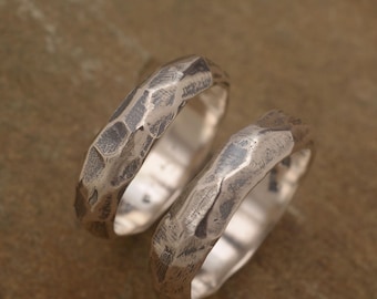May include: Two silver rings with a textured, hammered finish. The rings are displayed on a brown surface. The rings have a unique, irregular surface, catching the light and creating a rustic, handcrafted look. The rings are likely for a couple.