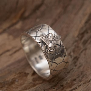 May include: A silver ring with a honeycomb pattern and a bee design. The ring is on a wooden surface.